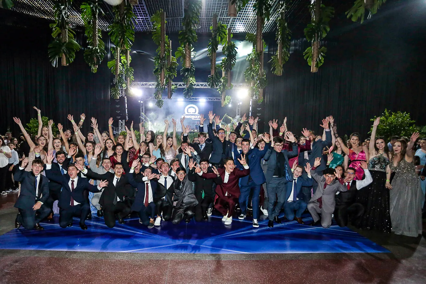 Turma de formandos celebrando reunida diante do palco iluminado.